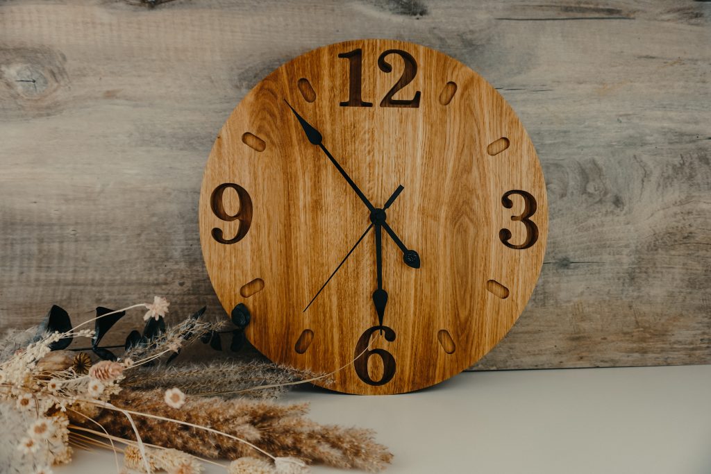 Hölzerne Wanduhr die auf einem Tisch steht und an einer grauen Wand lehnt.